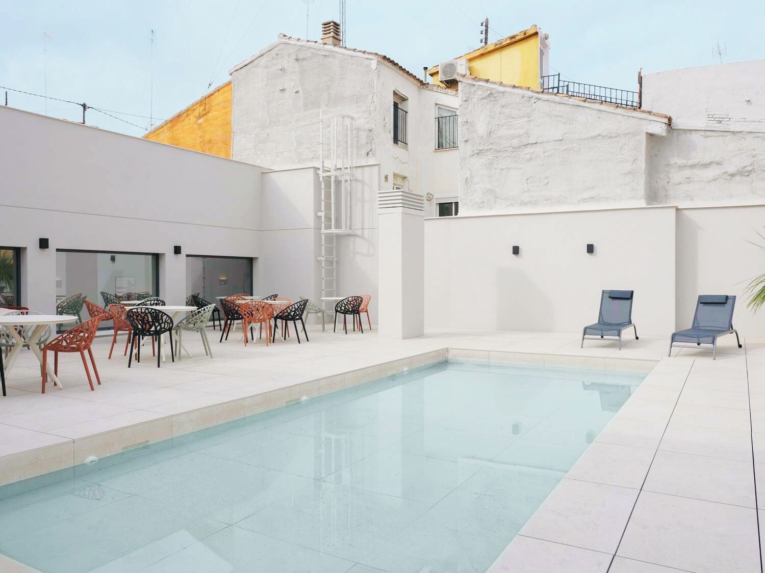 A modern outdoor pool area with a clear pool, surrounded by white walls and a yellow building. There are several tables and chairs for guests to enjoy the space.