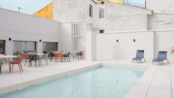 A modern outdoor pool area with a clear pool, surrounded by white walls and a yellow building. There are several tables and chairs for guests to enjoy the space.