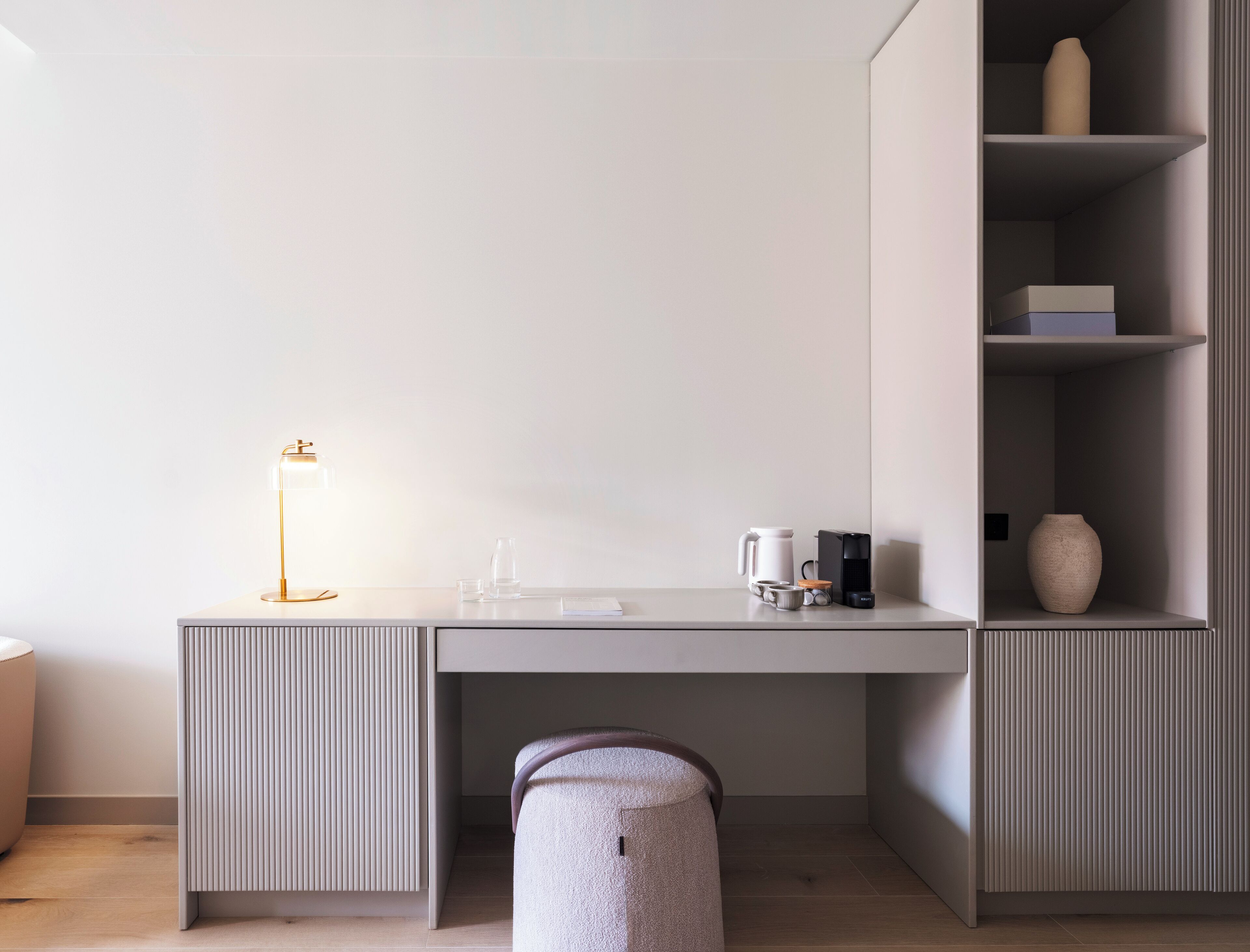 Una habitación moderna y minimalista con un escritorio gris, una lámpara, una cafetera y un florero en estantes.