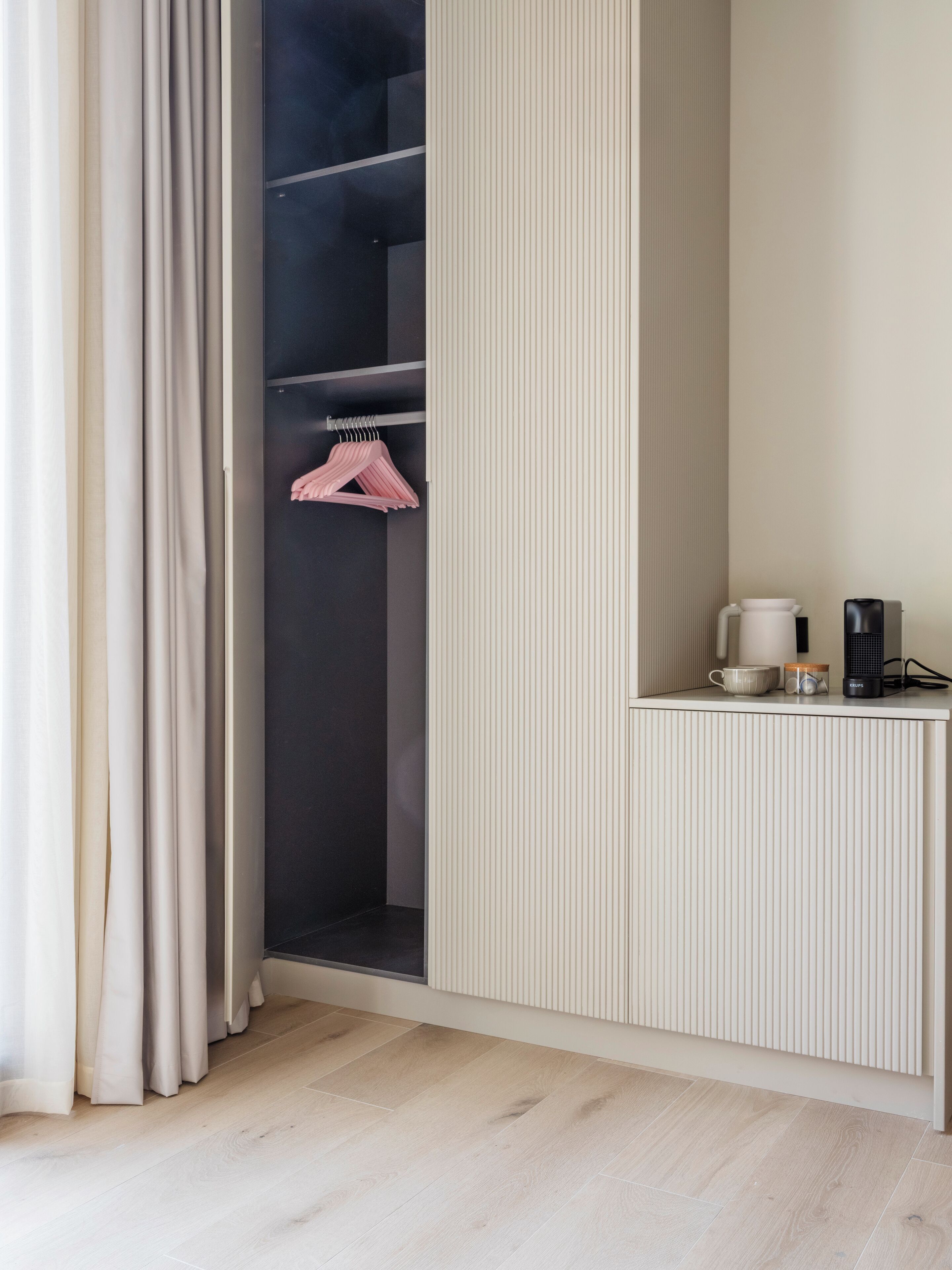 A modern room with a wooden floor, a built-in closet with hanging clothes, a radiator, and a small table with a coffee maker and a cup.