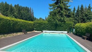 Une piscine rectangulaire entourée d’une terrasse en bois, le tout se fondant dans un décor de feuillus verdoyants et d’un ciel bleu azur.