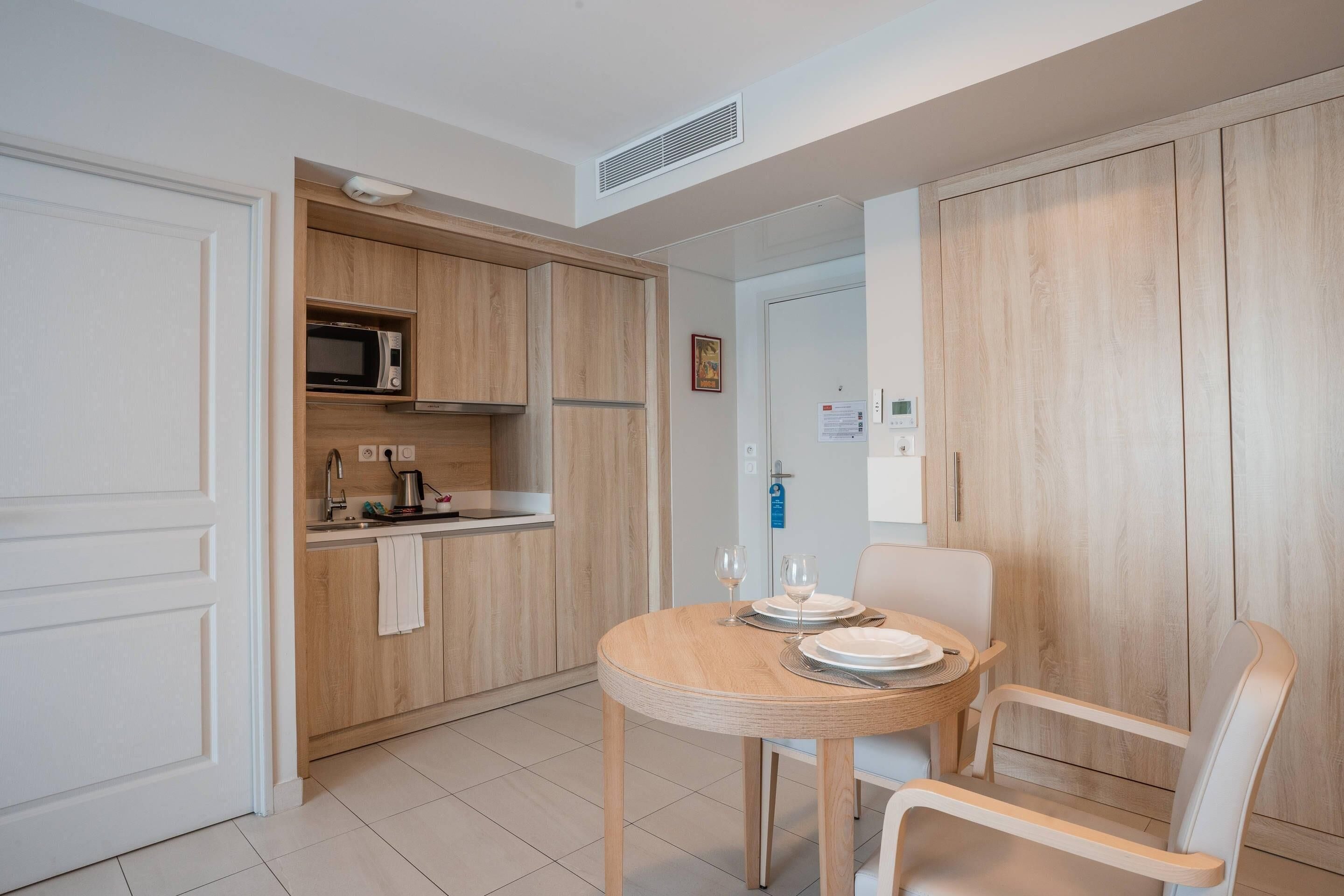 A compact kitchen with a microwave, sink, and wooden cabinets. There is a dining table with chairs and a small cabinet with a microwave above it.