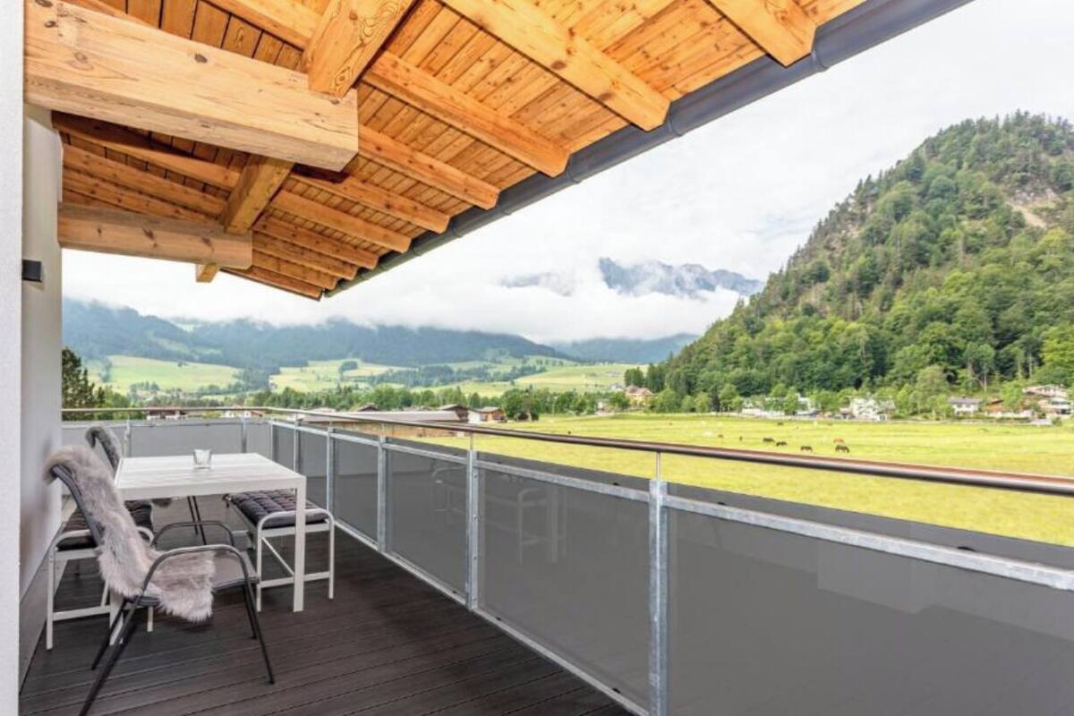 En balkong med ett träpanelat tak, ett bord och stolar, med utsikt över ett lantligt landskap med berg och åkrar.