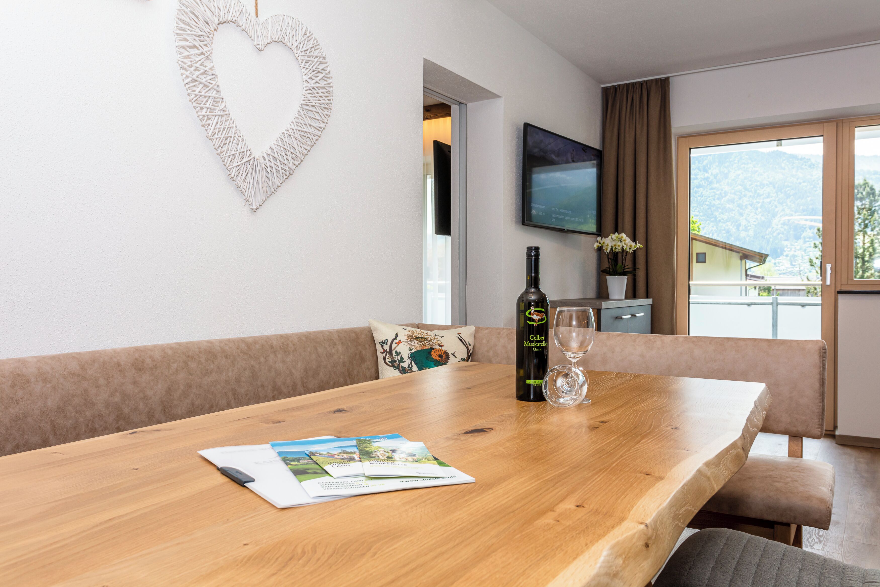 A wooden dining table with a magazine, a bottle of wine, and a glass. A heart-shaped decoration hangs on the wall, and a TV is mounted above a window with a view of mountains.