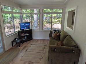 Una sala de estar con televisor sobre un mueble bajo, un sofá y ventanas amplias con vistas a árboles.