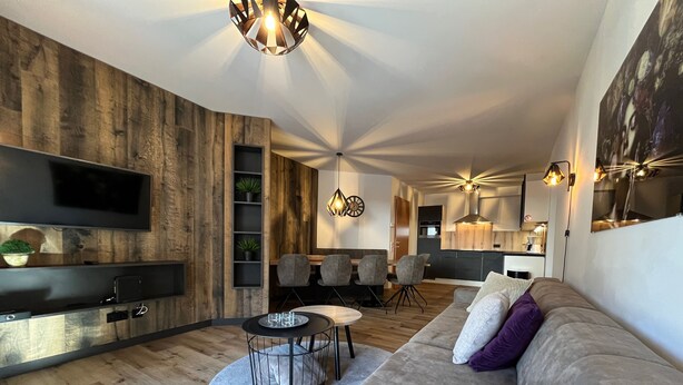 A modern living room with a wooden accent wall, a flat-screen TV, a dining area with chairs, and a kitchen in the background.