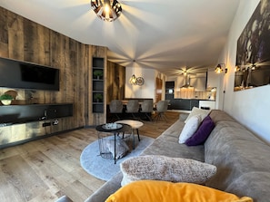 A modern living room with a wooden accent wall, a flat-screen TV, a dining area with chairs, and a kitchen in the background.