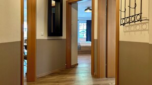 A hallway with wooden flooring, a mirror on the wall, and a door leading to a room with a bed and a window.