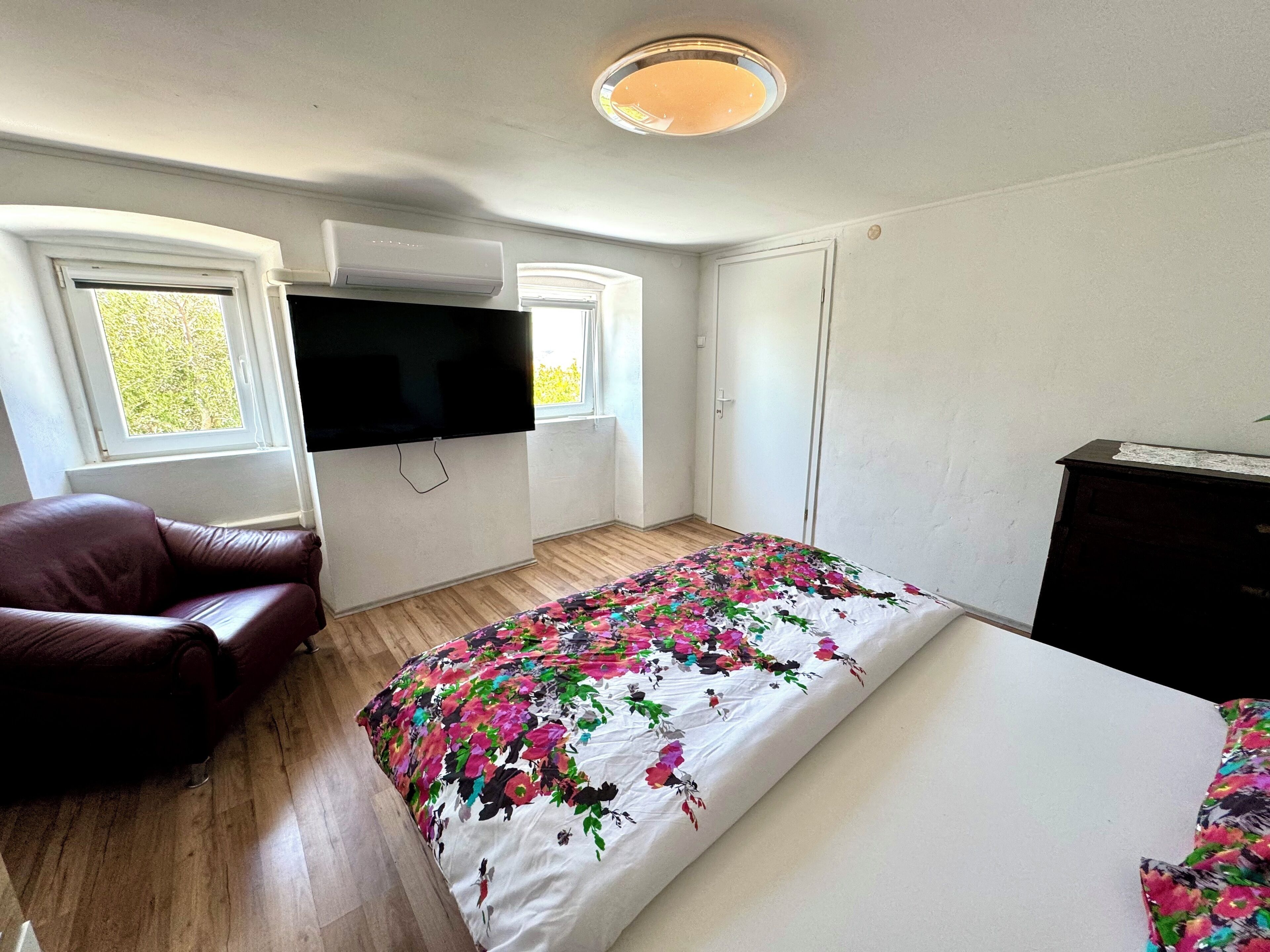 A bedroom with a bed, a TV mounted on the wall, a leather chair, and a window.