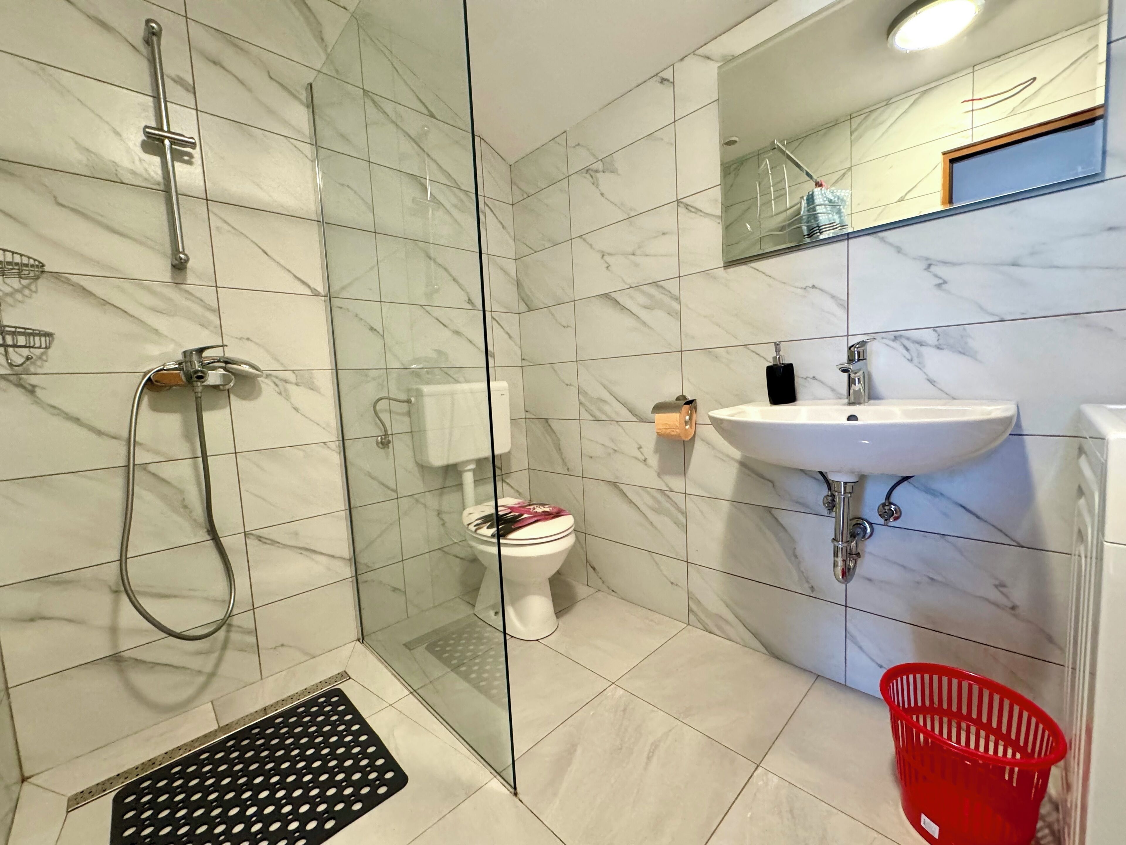 Een badkamer met een douchecabine van glas, een toilet, een wastafel en een rode mand.