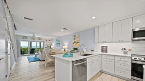 Een moderne keuken met witte kasten, een inbouwoven en een vaatwasser. Er is een eethoek met een blauw vloerkleed en een witte stoel.