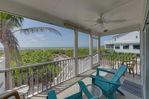 A covered outdoor area with a view of the ocean, featuring a ceiling fan, a table, chairs, and a railing.