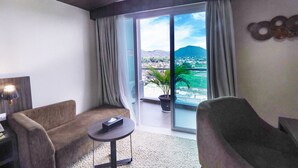 A modern hotel room with a large sliding glass door leading to a balcony with a view of a cityscape and mountains. There are two armchairs and a small round table with a remote control on it. - Labersa Hotel & Convention Centre Toba (Balige)