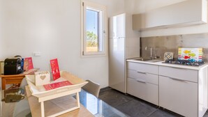 Une cuisine compacte équipée d’un réfrigérateur, d’une cuisinière à gaz et d’un évier. On y trouve une petite table avec une chaise, ainsi qu’une fenêtre qui laisse entrer la lumière naturelle.