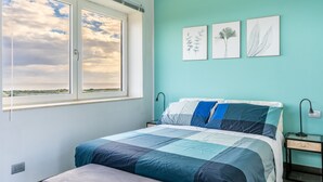 Une chambre avec un grand lit, une fenêtre donnant sur un beau paysage et trois illustrations de plantes accrochées au mur.