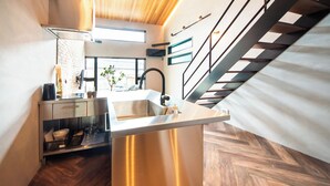 A modern kitchen with a stainless steel island, wooden herringboard flooring, and a staircase with black railings. - Private House Hibiki (Nara)