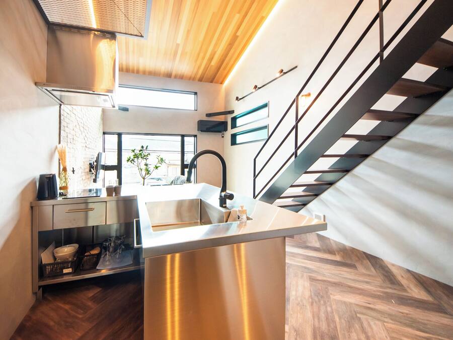 Una cocina moderna con una isla de acero inoxidable, piso de tablones de madera y una escalera con barandales negros.