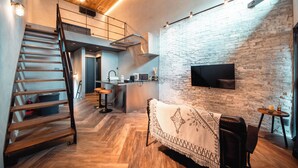 A modern living space with a wooden staircase, a sofa, a TV mounted on a brick wall, and a kitchen area in the background. - Private House Hibiki (Nara)