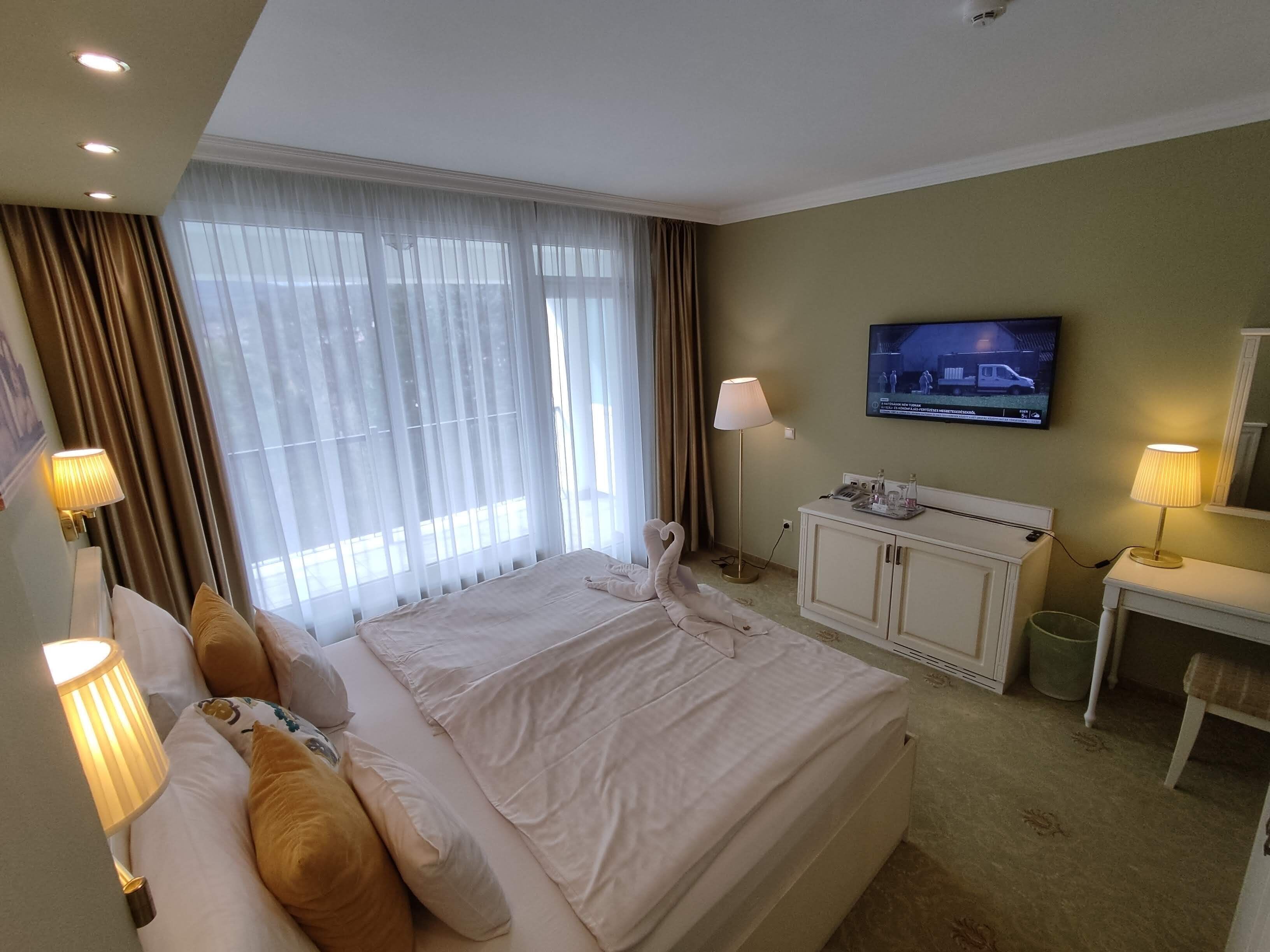 A hotel room with a large bed, a television on the wall, a desk with a lamp, and a window with curtains.
