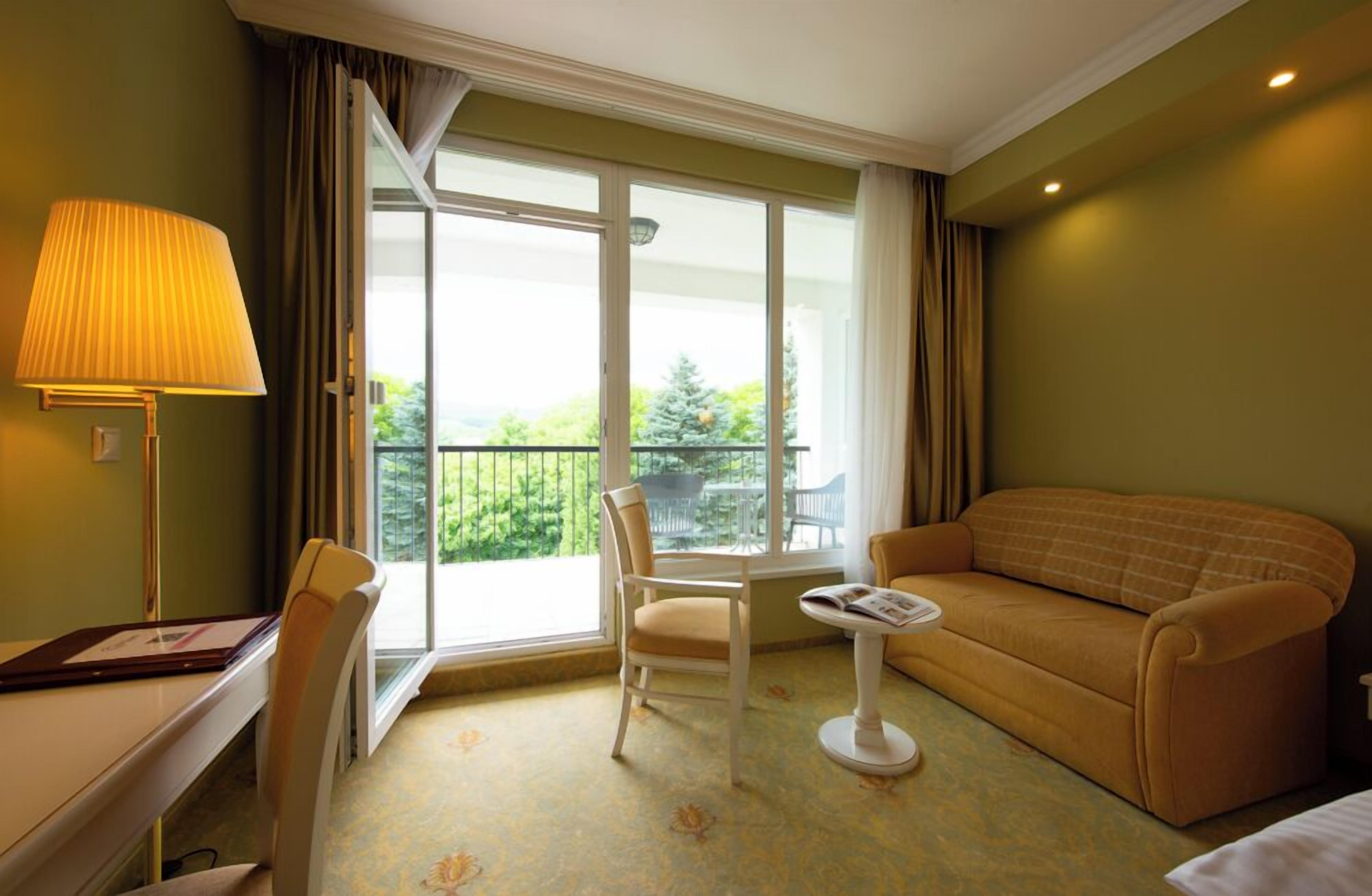 A hotel room with a sofa, a chair, a small table, a lamp, and a large window offering a view of the outdoors.