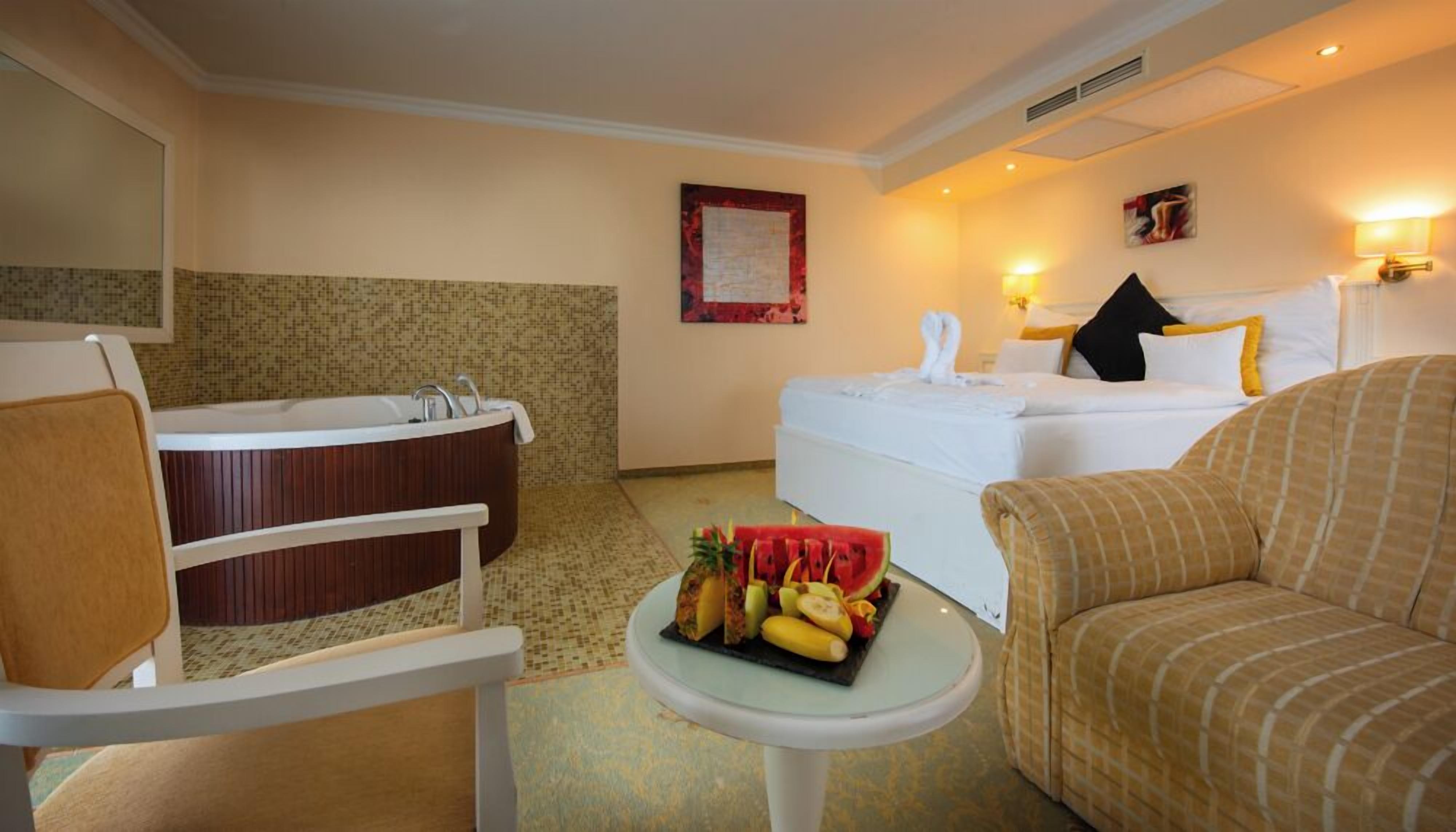 A hotel room with a bathtub, a bed, a sofa, and a small table with fruit.