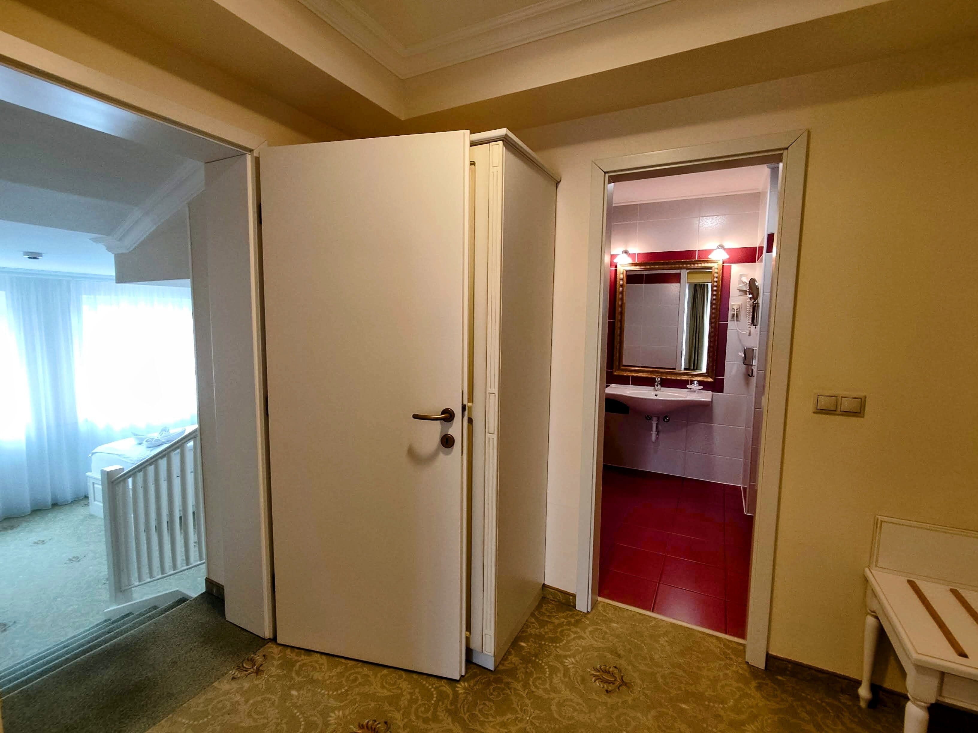 Habitación de hotel con puerta cerrada, una mesita de noche pequeña, baño con lavamanos y espejo, y suelo de baldosas rojas.