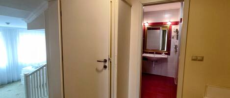 Habitación de hotel con puerta cerrada, una mesita de noche pequeña, baño con lavamanos y espejo, y suelo de baldosas rojas.