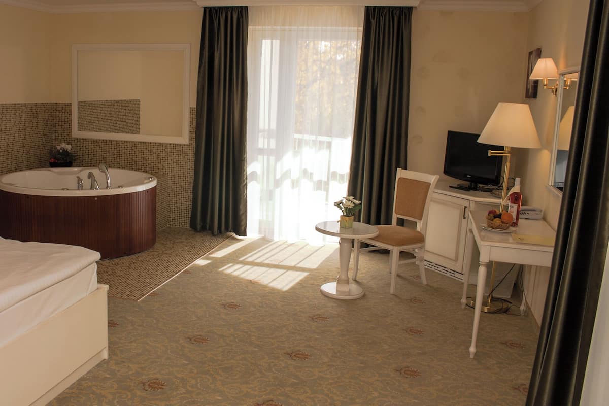 Quarto de hotel com uma banheira grande, cama, escrivaninha com cadeira, televisão e janela com cortinas.