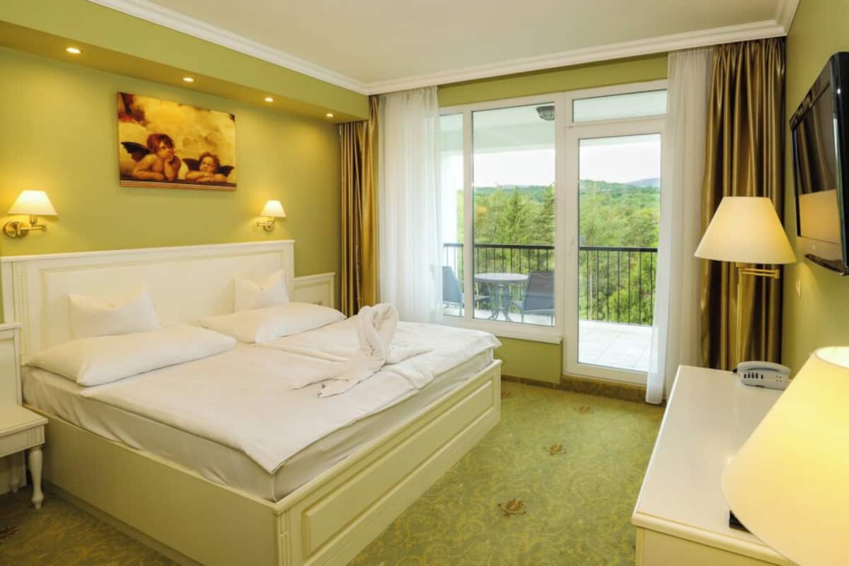 Quarto de hotel com uma cama grande, duas luminárias de cabeceira, uma televisão e uma varanda com vista para o verde.