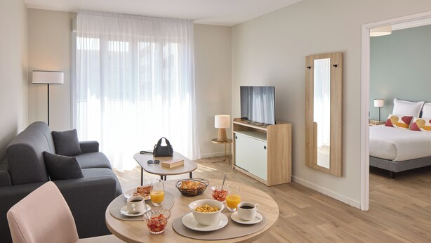 A modern hotel room with a sofa, a dining table with breakfast, a TV, and a bed. - Quality Aparthotel Reims - Saint-Thomas (Reims)