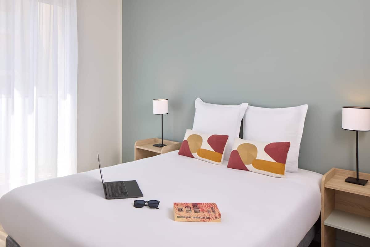 A neatly made bed with a laptop, sunglasses, and a book on a hotel room with a light blue wall and white curtains.