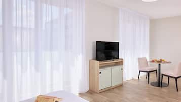 A modern hotel room with a bed, a TV on a wooden cabinet, a dining table with chairs, and sheer curtains.