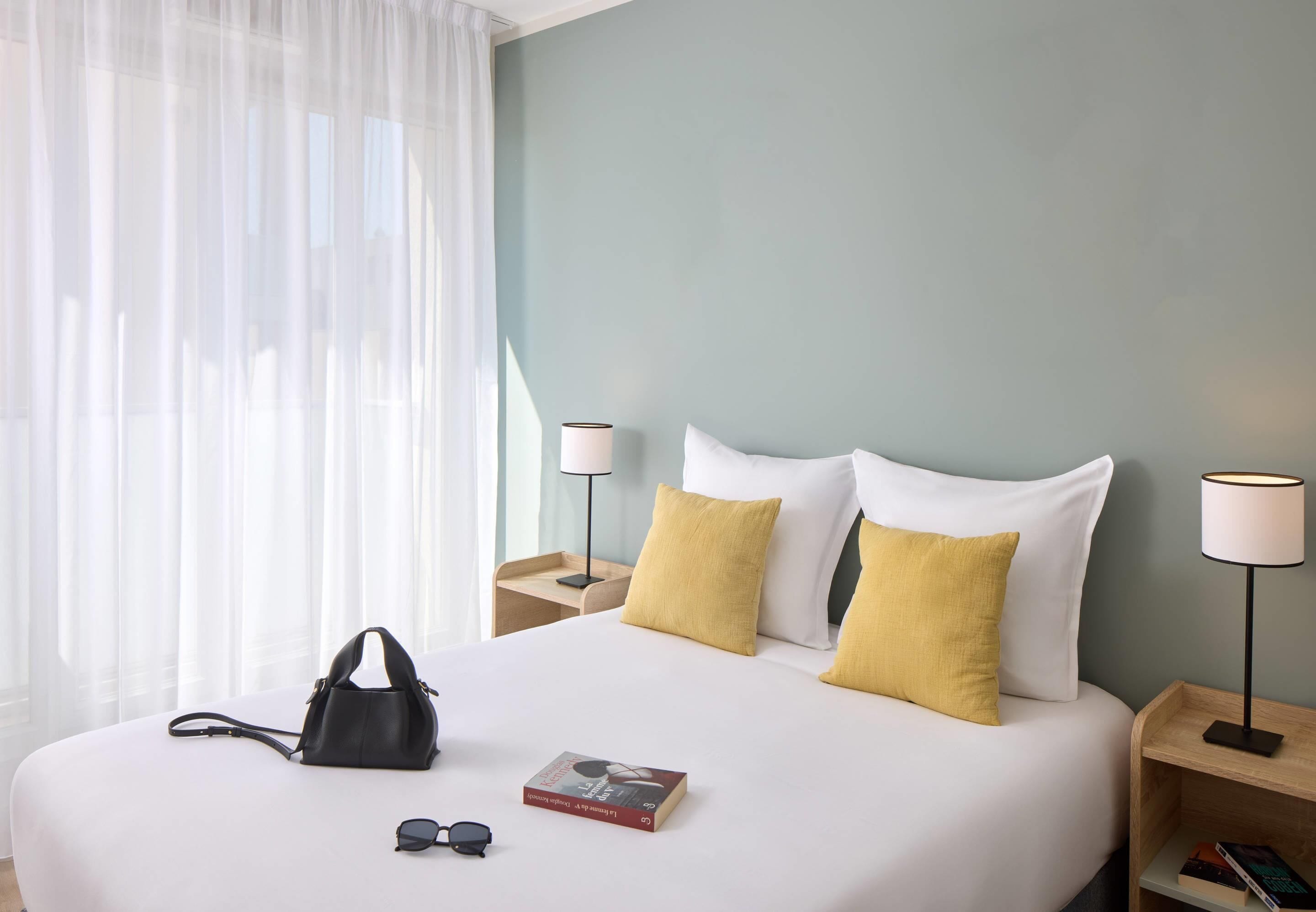 A neatly made bed with white and yellow pillows, a black handbag, and a book on the bed. There are two bedside tables with lamps and a window with sheer curtains.