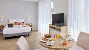 A hotel room with a bed, bedside tables, a TV, breakfast table with food and drinks, and a window with curtains. - Quality Aparthotel Reims - Saint-Thomas (Reims)
