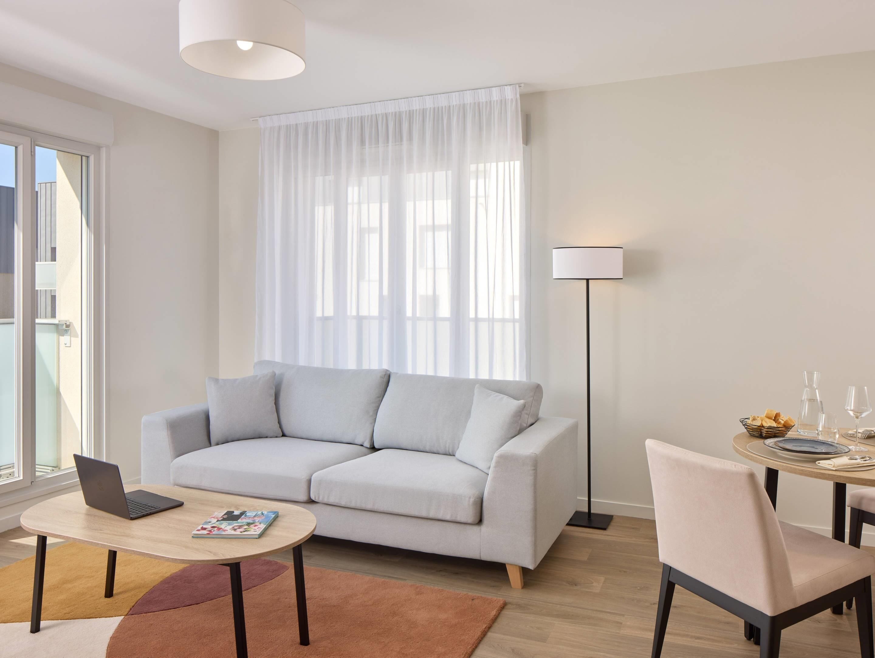 A modern living room with a grey sofa, a wooden coffee table, a laptop, a floor lamp, and a dining area with a table and chairs.