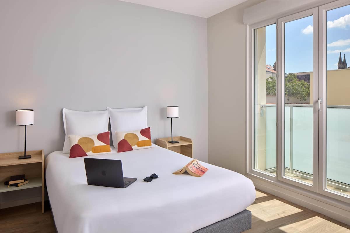 A modern hotel room with a large bed, two bedside tables, a laptop, sunglasses, and a book.