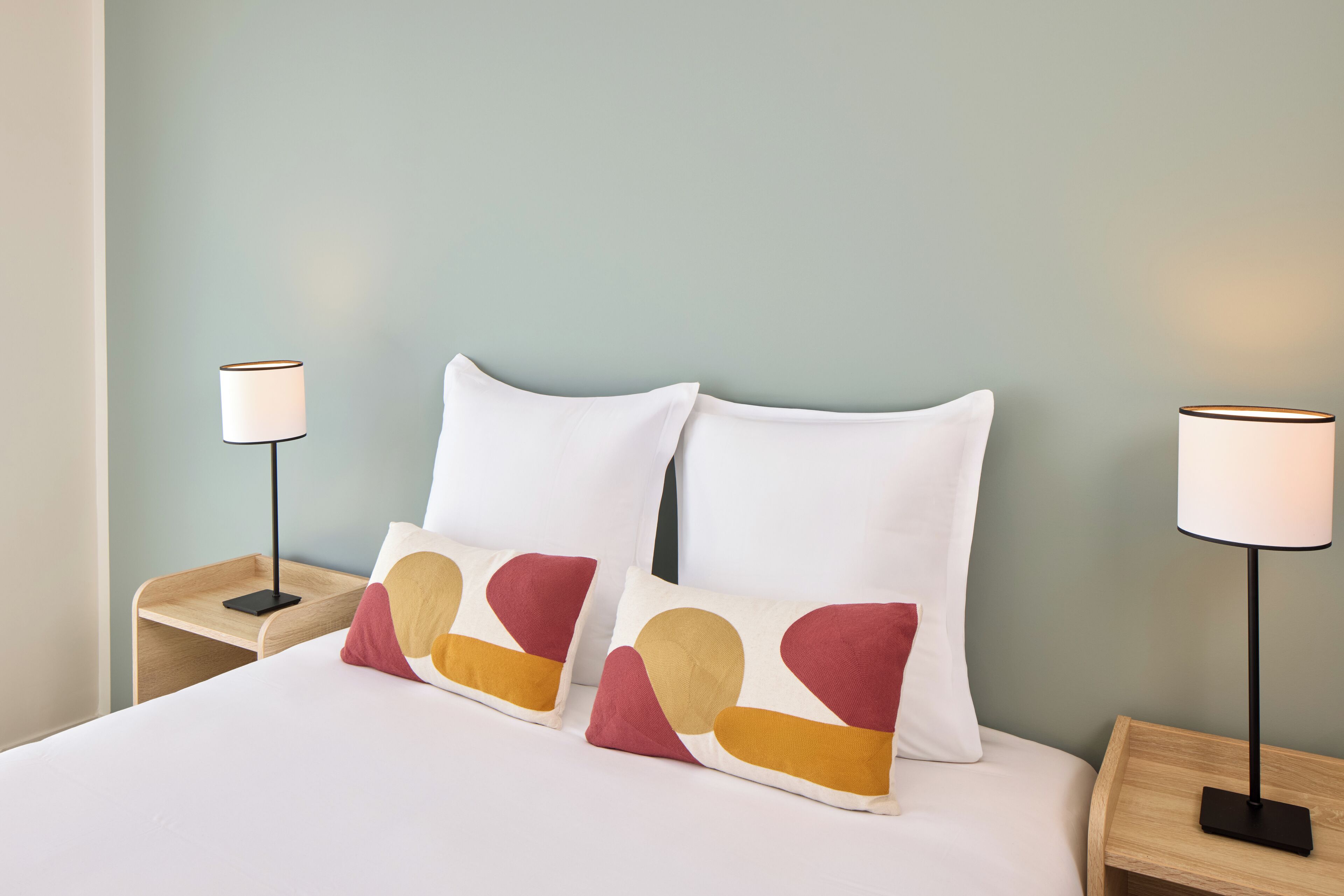 A neatly made bed with white bedding and two decorative pillows with abstract patterns. There are two bedside tables, each with a lamp. The walls are painted in a light teal color.