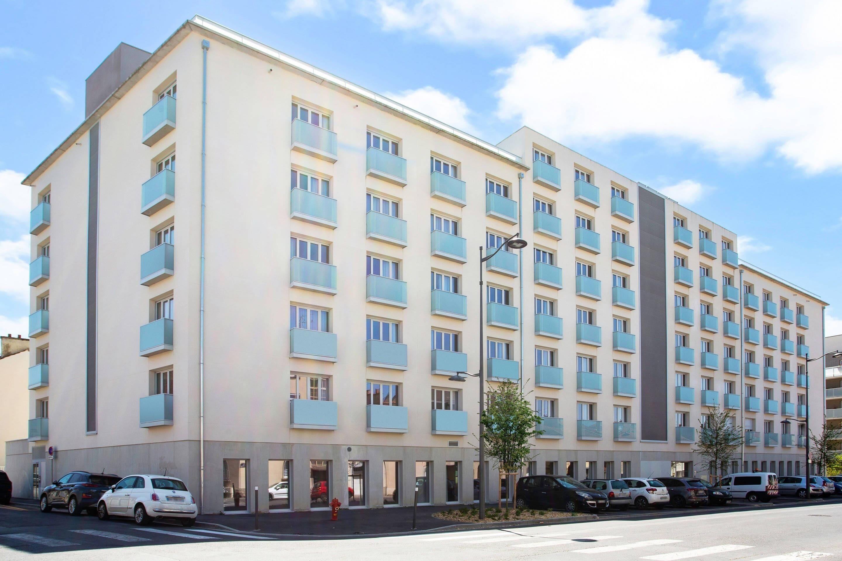 Ett hotell med flera våningar, en beige fasad, balkonger och en parkeringsplats med flera bilar.