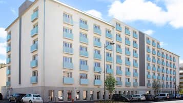 A multi-story hotel with a beige facade, balconies, and a parking area with several cars.