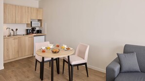 Een compacte keuken met houten kastjes, een magnetron en een koffiezetapparaat. Er is een eettafel met stoelen en een grijze bank.