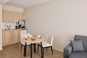 A compact kitchen with wooden cabinets, a microwave, and a coffee maker. There is a dining table with chairs and a grey sofa. - Quality Aparthotel Reims - Saint-Thomas (Reims)