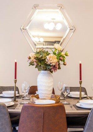Ein Esstisch für vier Personen, gedeckt mit weißen Blumen in einer Vase, roten Kerzen und einem sechseckigen Spiegel darüber.