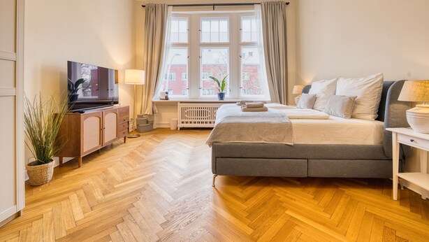 A bedroom with a bed, a TV, a plant, and a window with curtains. - Premium Suite in West Berlin, Sleeps 4, Family & Group Friendly, 70m to Metro (Berlin)