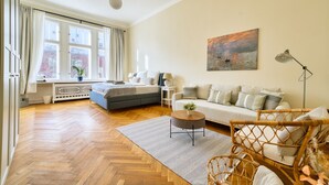 A spacious living room with a herringbone wooden floor, a grey sofa, a blue sofa, a wicker chair, and a painting on the wall. - Premium Suite in West Berlin, Sleeps 4, Family & Group Friendly, 70m to Metro (Berlin)