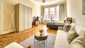 A modern living room with a sofa, a coffee table, and a TV. There is a wardrobe, a bed, and a window with curtains. - Premium Suite in West Berlin, Sleeps 4, Family & Group Friendly, 70m to Metro (Berlin)
