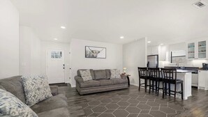 Un salon moderne avec un canapé gris, un coin repas avec une petite table et des chaises, et une cuisine avec des meubles blancs et un réfrigérateur.