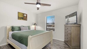 Une chambre à coucher comprenant un lit, une commode, un ventilateur de plafond et deux tableaux accrochés au mur.