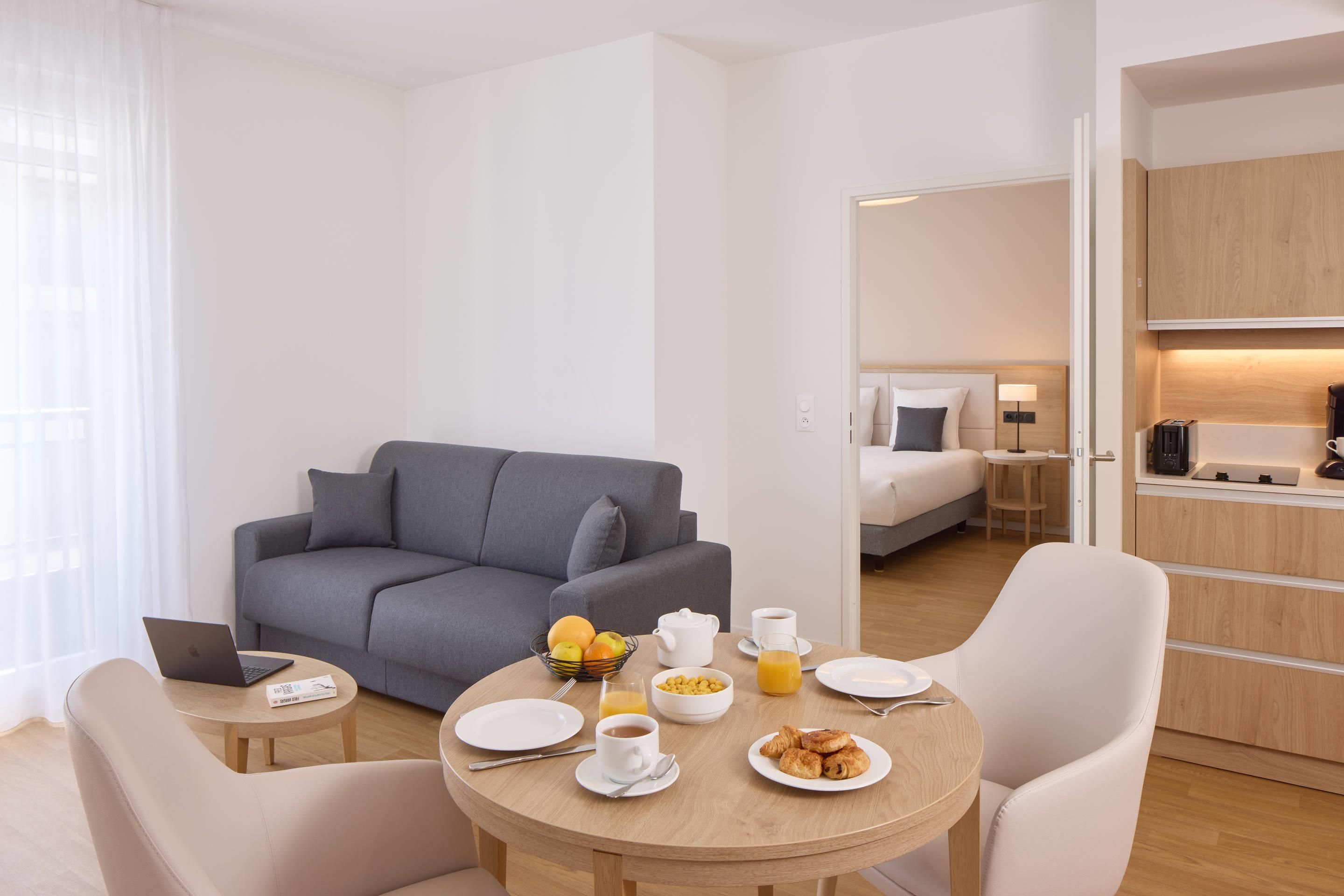 Et moderne hotelværelse med en grå sofa, et rundt spisebord med morgenmadsretter, en bærbar computer og en seng i baggrunden.
