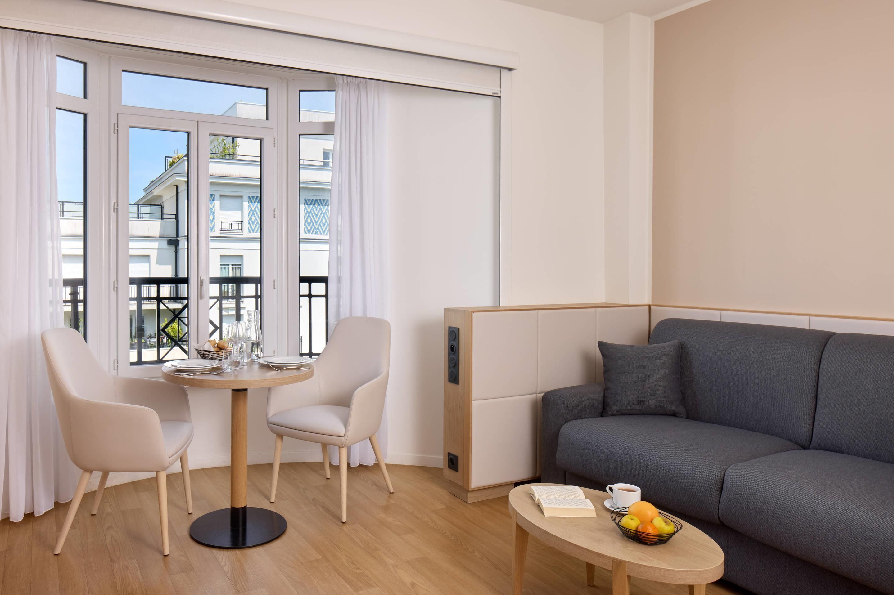 A modern living room with a grey sofa, a round table with a fruit bowl, and two chairs.