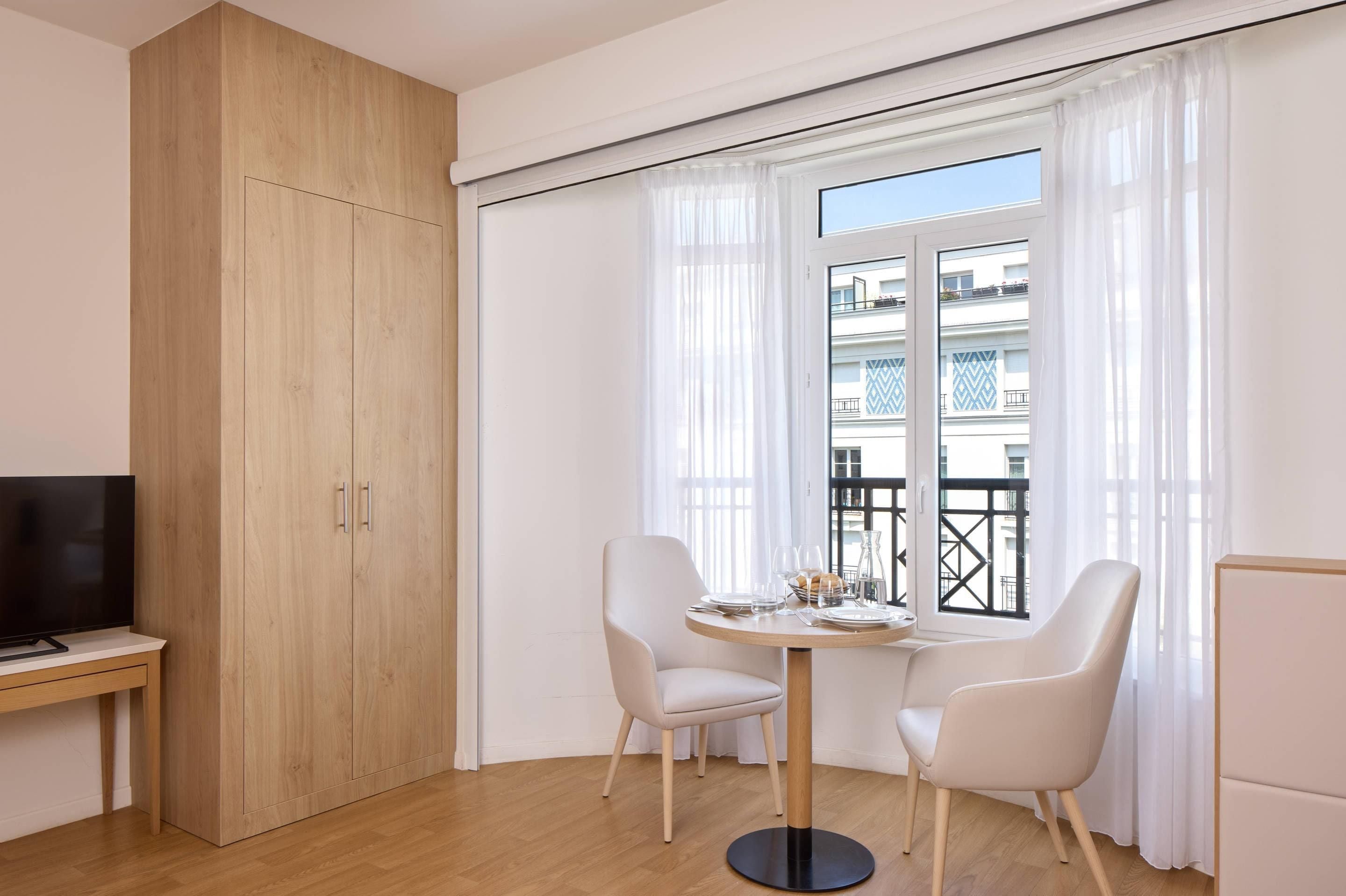 A modern living room with a wooden wardrobe, a flat-screen TV, a round dining table with two chairs, and a large window with sheer curtains.